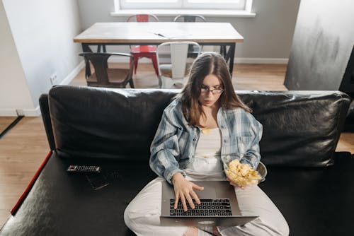 Woman Sitting On Sofa Using Her Laptop With Bowl Of Junk Food 
