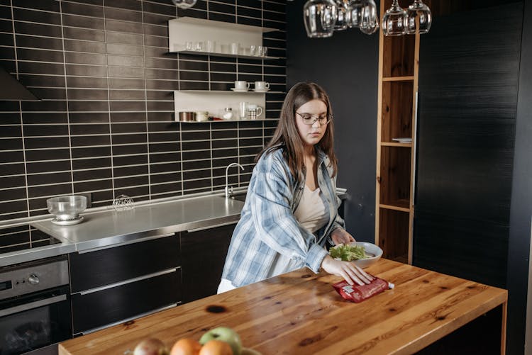 Woman In Blue Checked Shirt And Eyeglasses Putting Food On A Kitchen Table