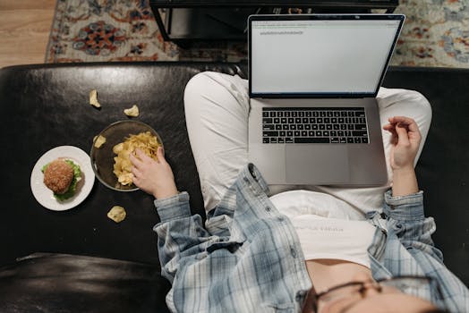 A person using a laptop while snacking on chips and a burger, creating a casual work-from-home vibe.