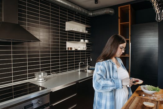 A woman prepares a meal in a stylish kitchen with dark tiling and stainless steel appliances.