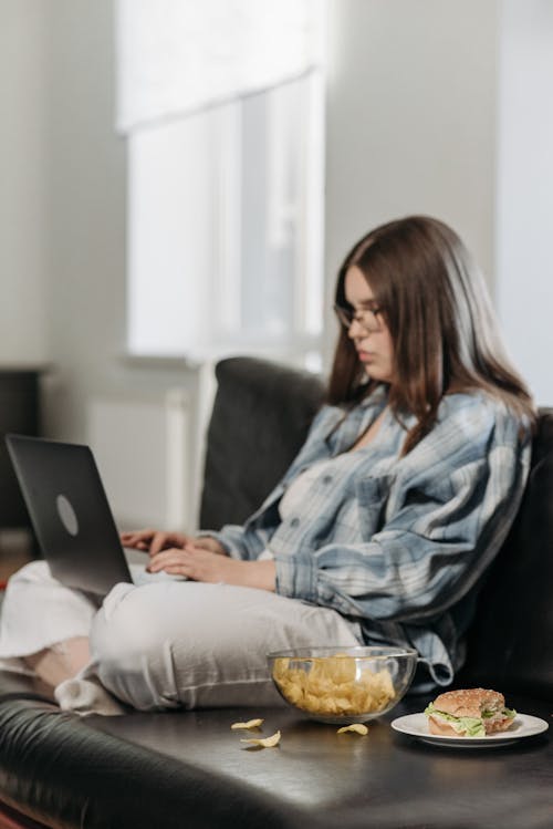 Woman in Blue and White Plaid Shirt Using Her Laptop While Sitting On Sofa Beside A Bowl Of Food