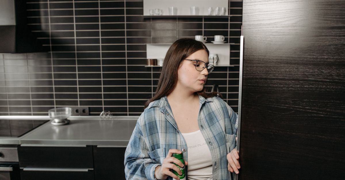Exhausted woman standing in front of open refrigerator late at night, holding a snack