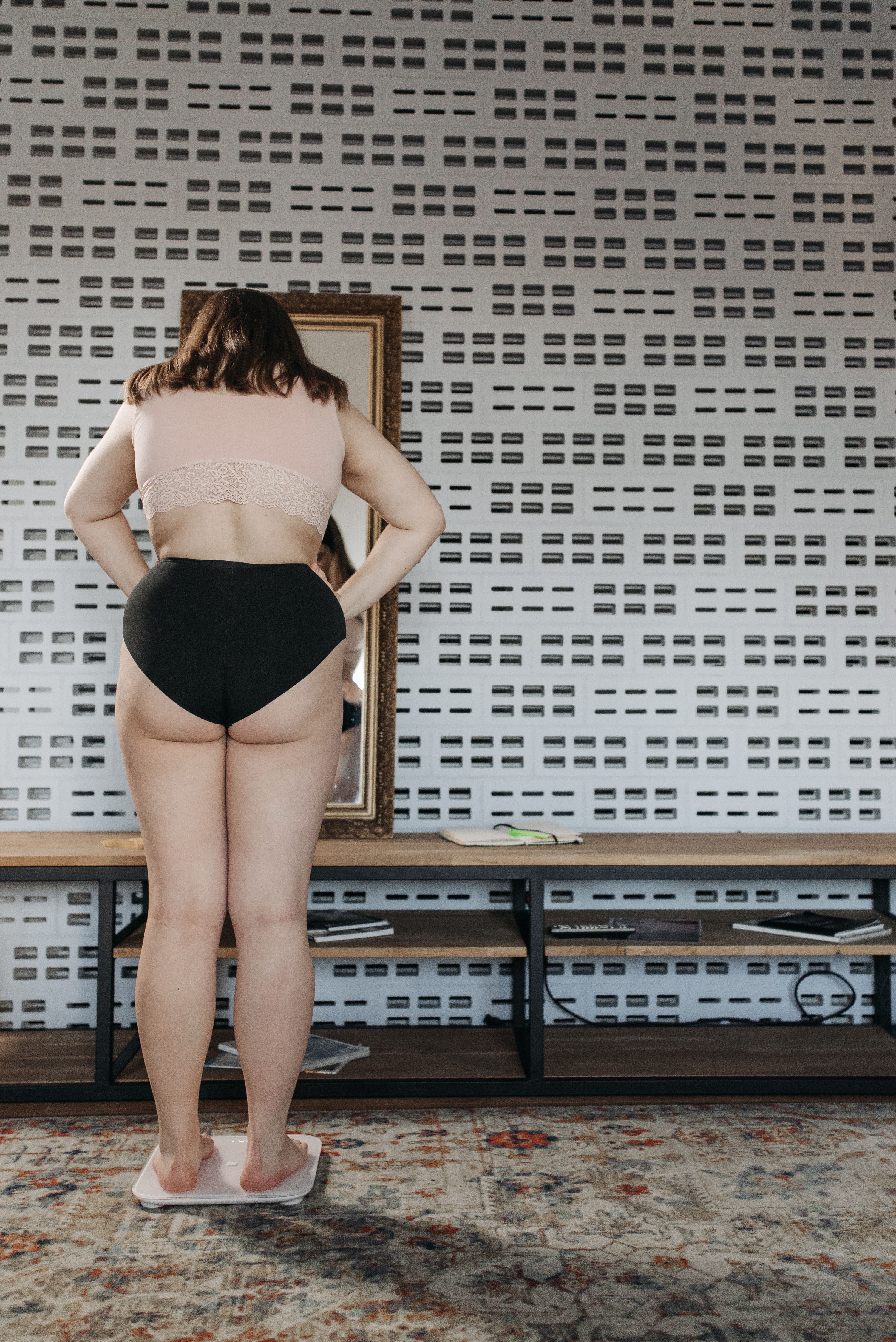 Plus-size woman measuring weight on scale in modern room with patterned wall.