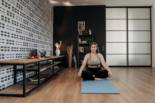 A woman practices meditation, sitting cross-legged on a yoga mat indoors, embodying relaxation and wellness.