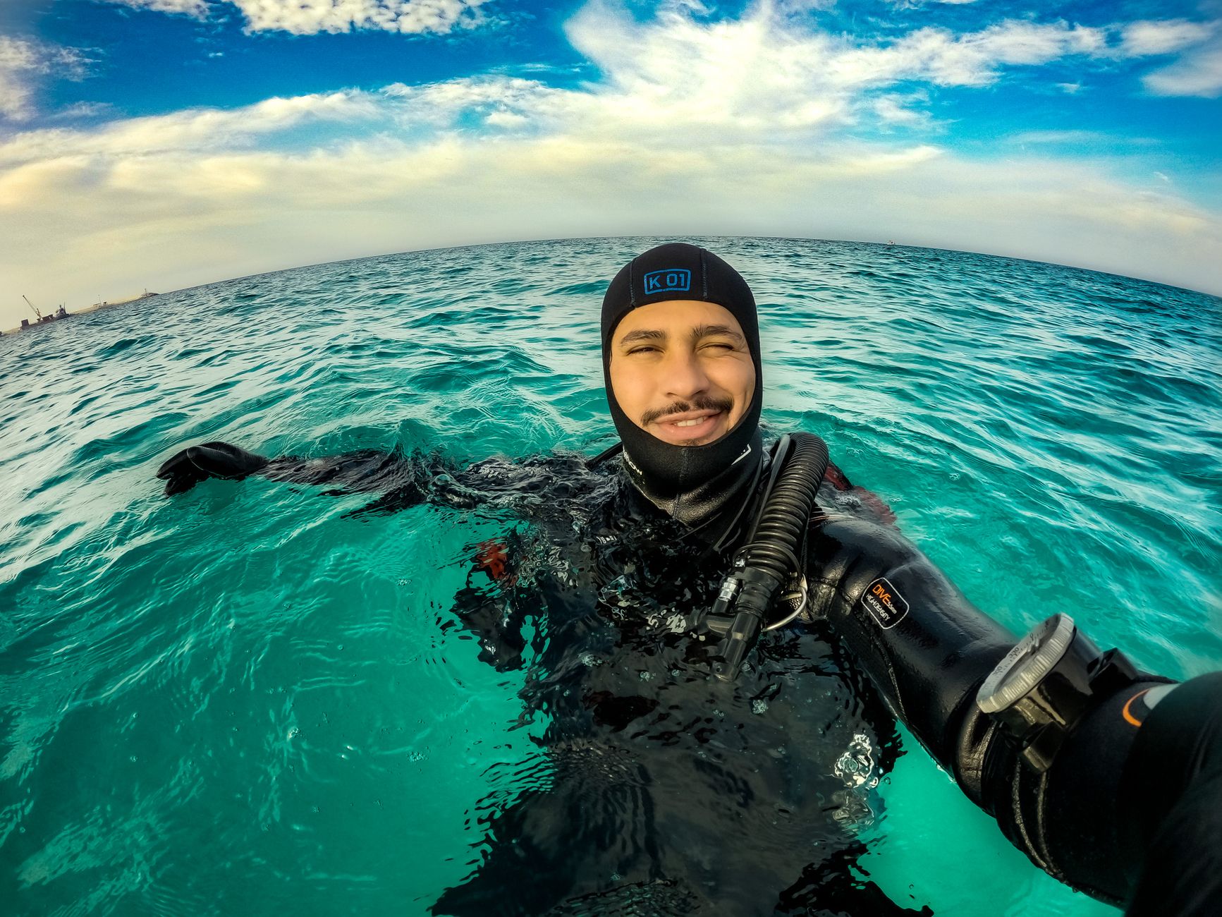 A scuba diver in a wetsuit smiles while taking a selfie in the clear blue sea under a partly cloudy sky. โ photo by Kevin Galeano / Pexels