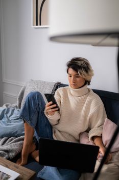 Asian woman in cozy sweater using smartphone and laptop on sofa indoors.