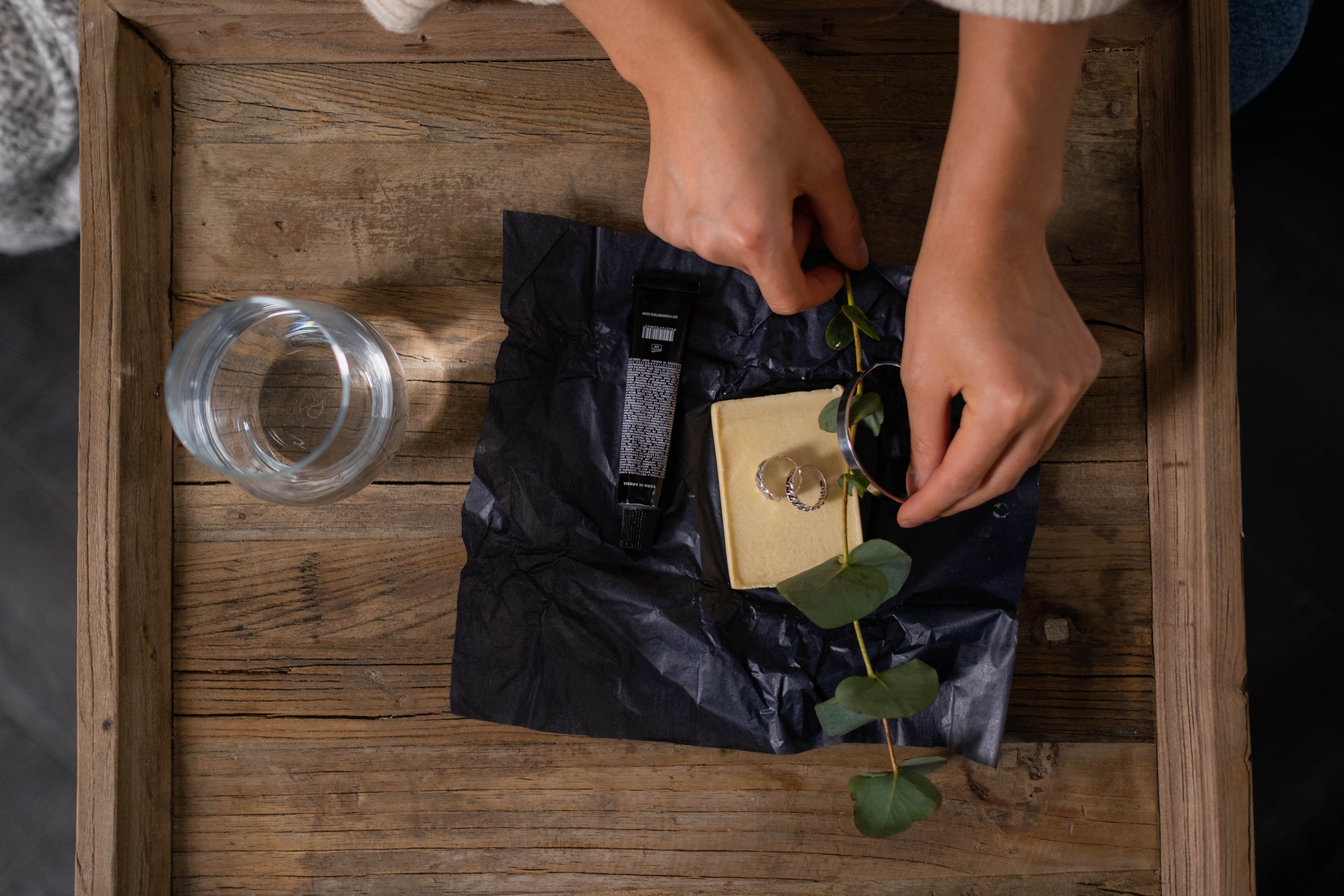 Ett elegant flat lay med ringar, eucalyptus och ett glas på en träyta, fångat i en varm inomhusmiljö.