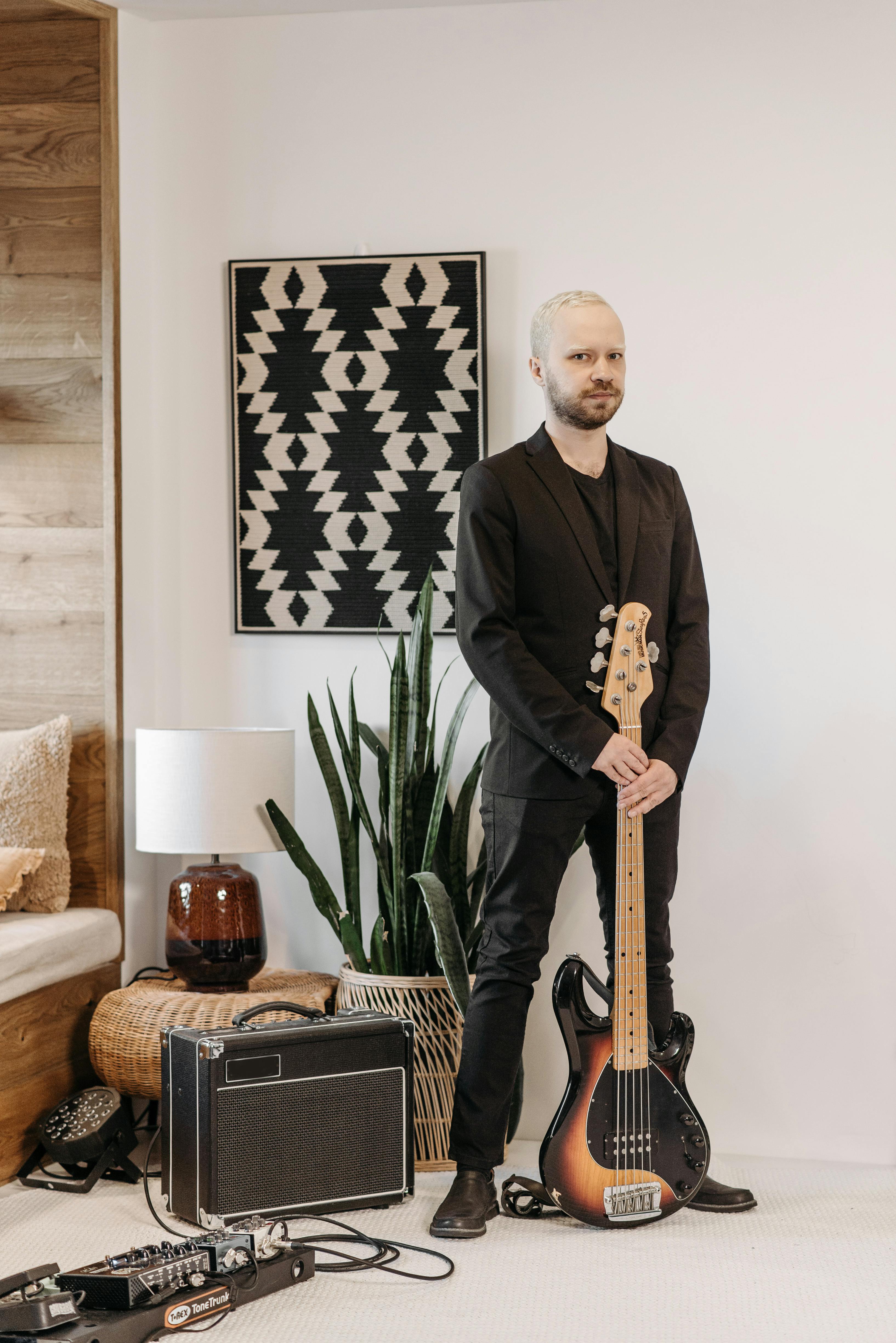A man in a black suit stands indoors with an electric guitar, amplifier, and a stylish decor.