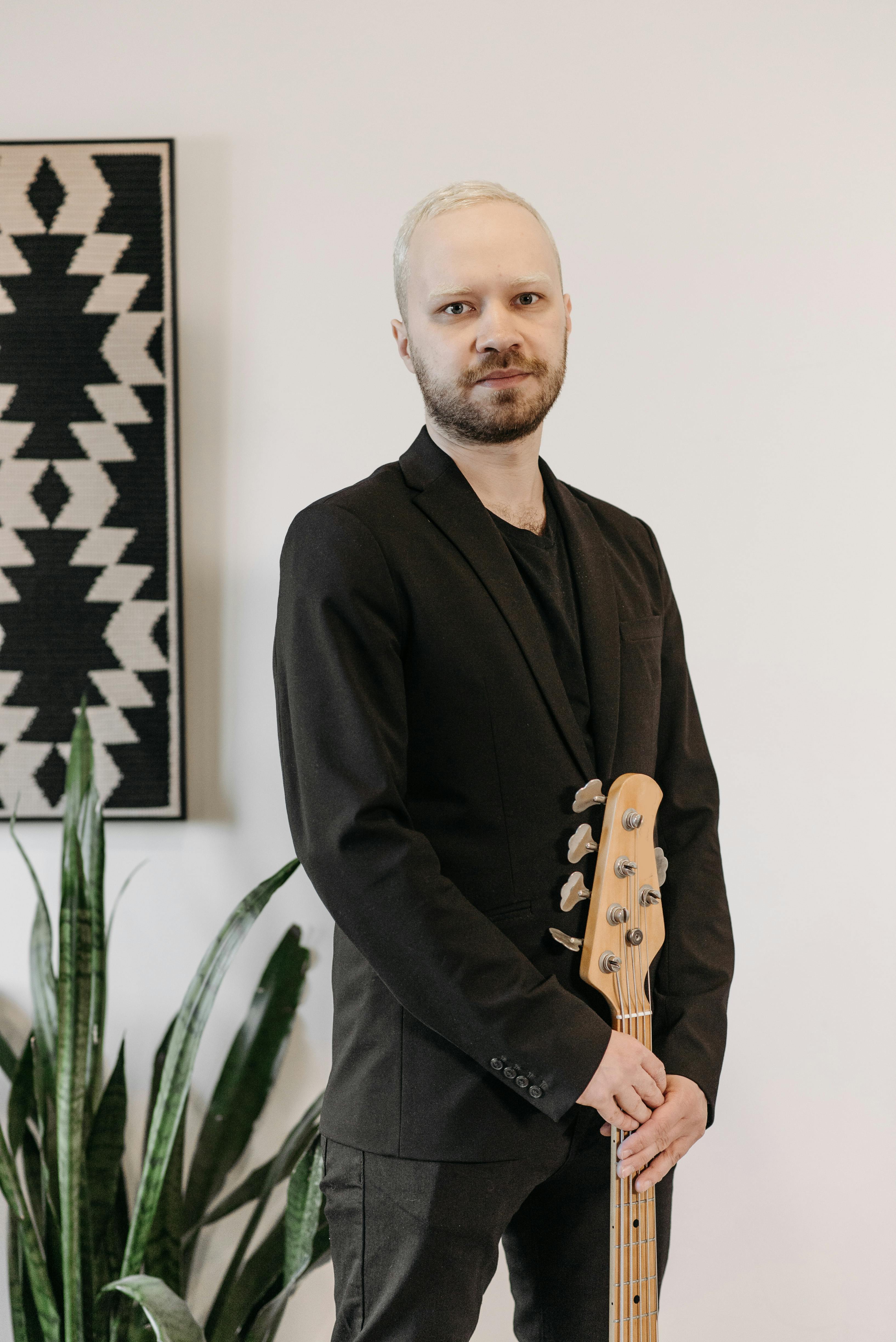 A musician in a black suit holding a bass guitar, standing indoors. Artistic and modern vibe.