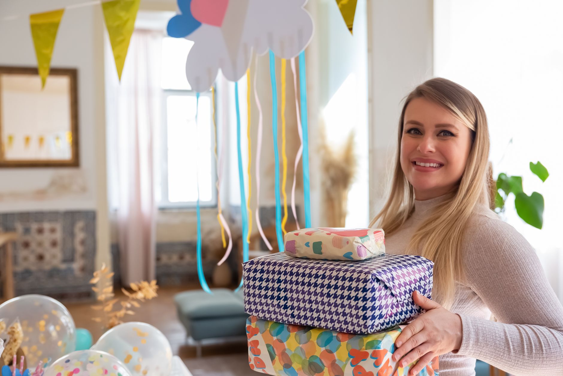 Happy woman holding colorful gift boxes at a festive baby shower party indoors.