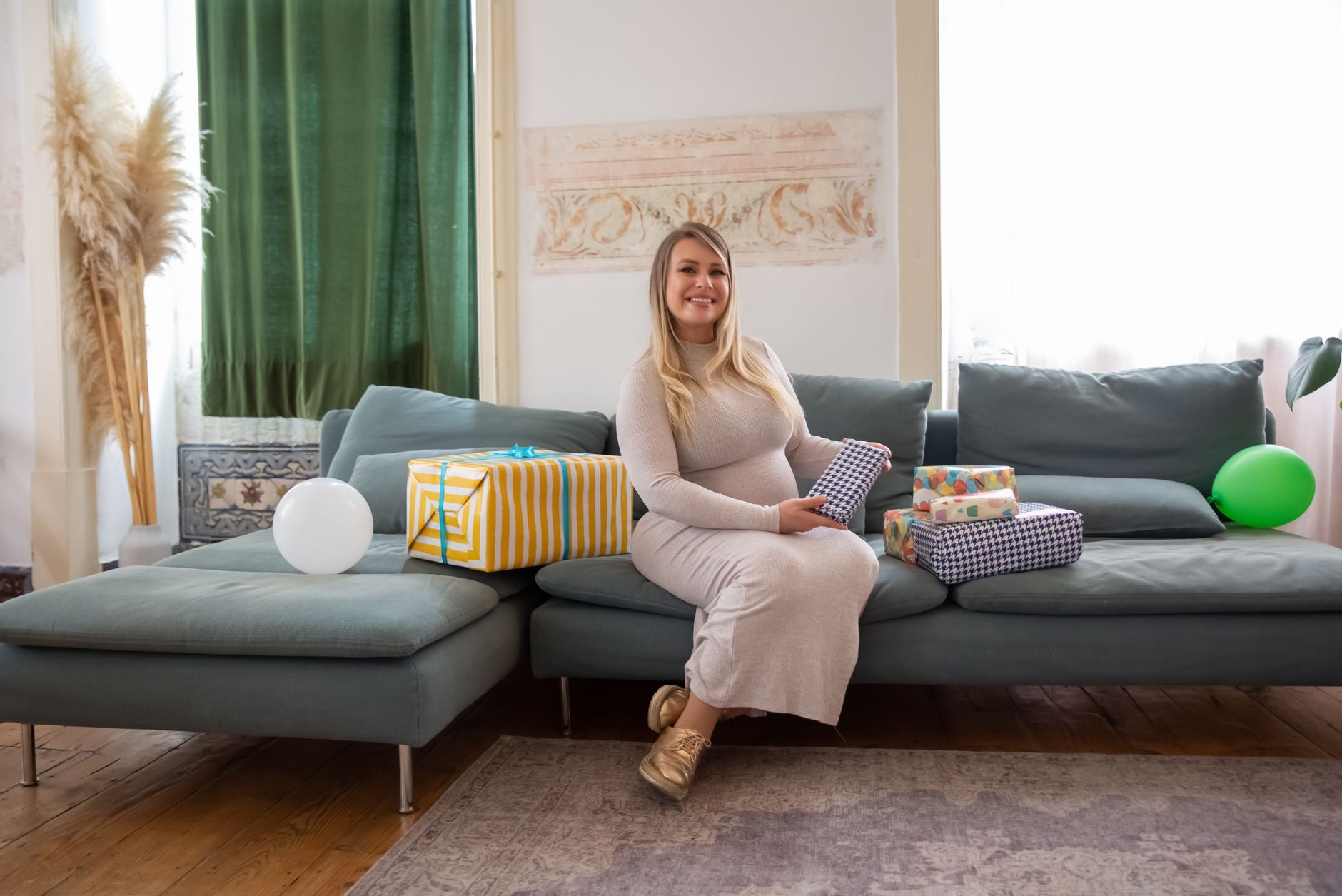 A joyful pregnant woman seated on a sofa, celebrating a baby shower with gifts in a cozy home setting.