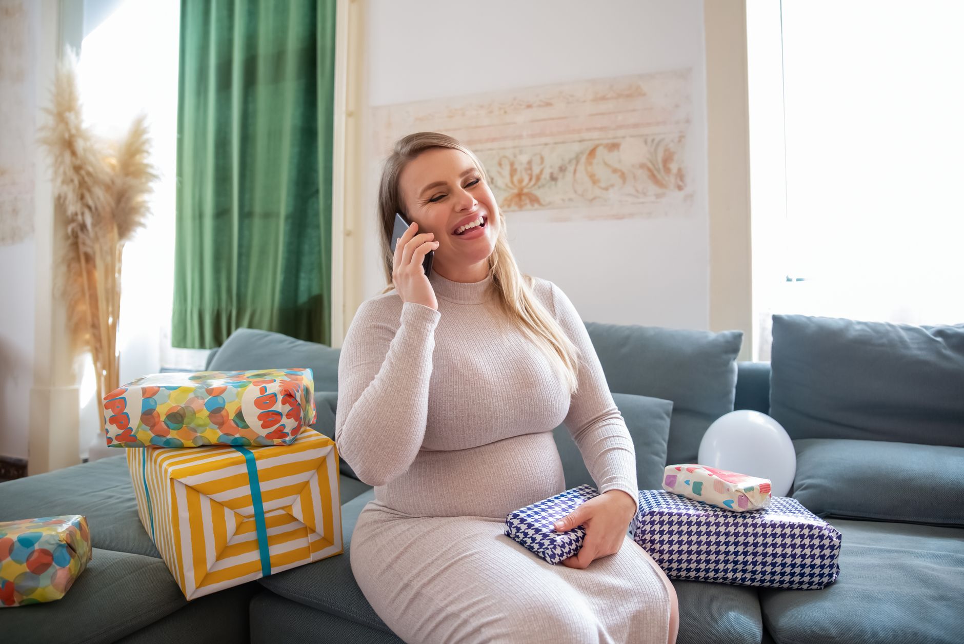 Cheerful pregnant woman on phone surrounded by gifts, enjoying a happy moment indoors.