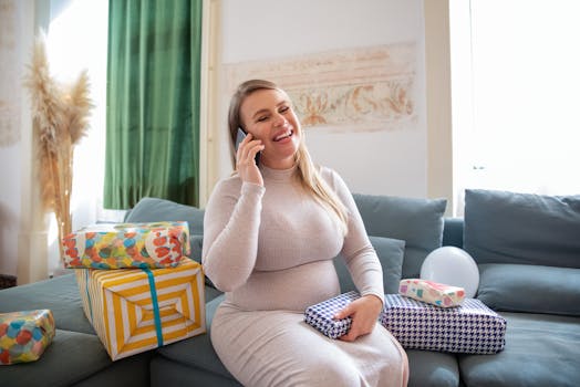 Cheerful pregnant woman on phone surrounded by gifts, enjoying a happy moment indoors.