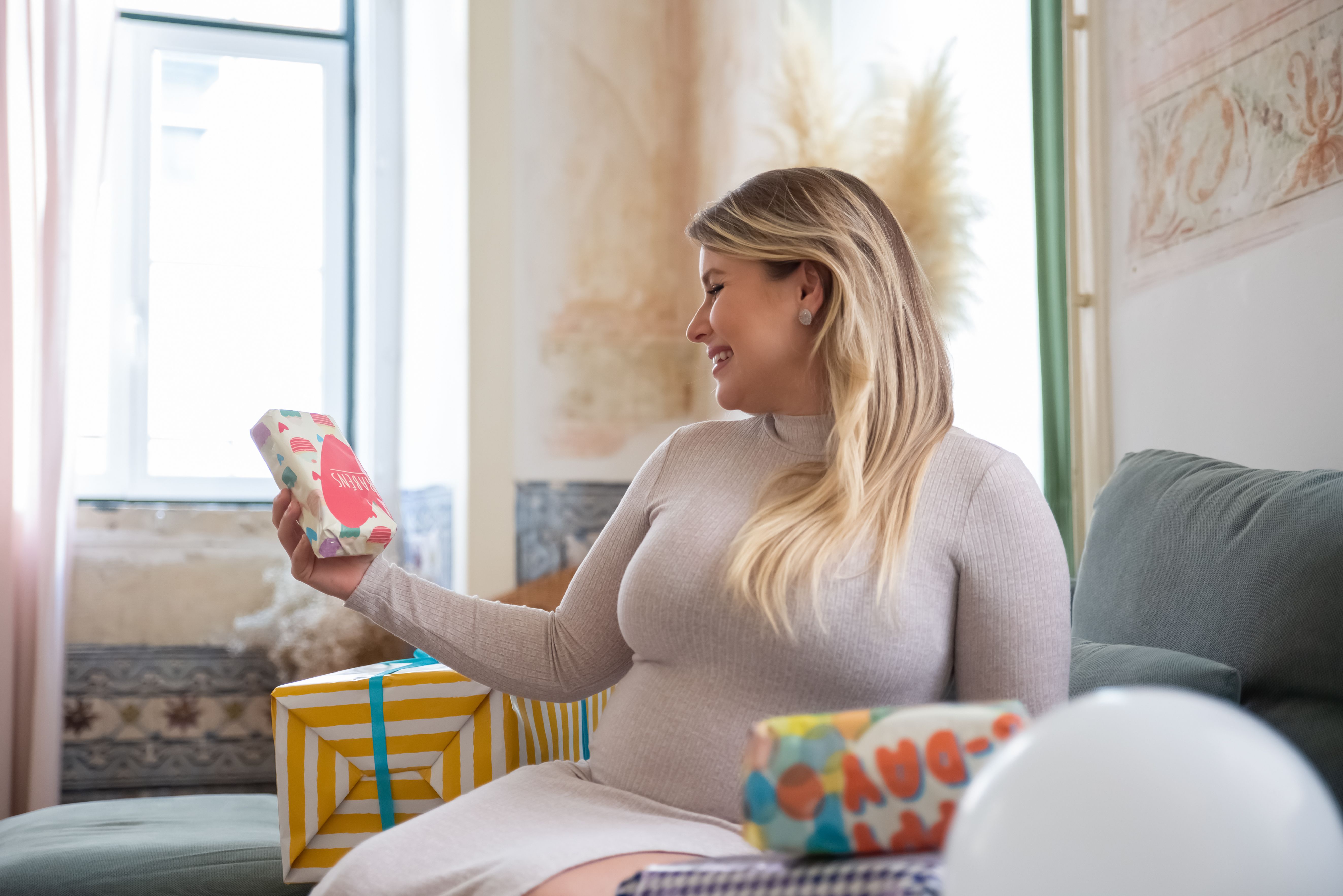 Pregnant woman smiling while holding gifts at a cozy indoor baby shower.