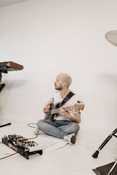 A bearded man plays an electric bass guitar in a minimalist studio, surrounded by music equipment.