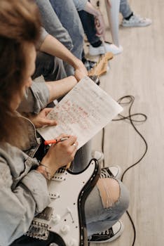 Musician writing song lyrics on manuscript while holding an electric guitar, creating music indoors.