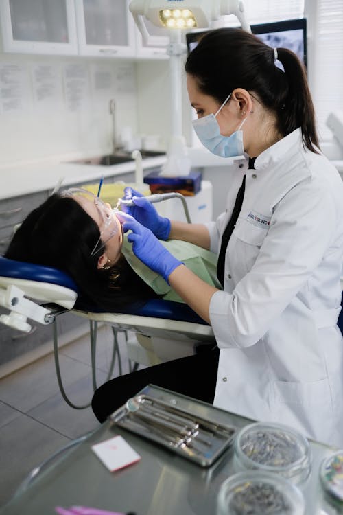 A Woman in White Coat Cleaning the Teeth of Her Patient