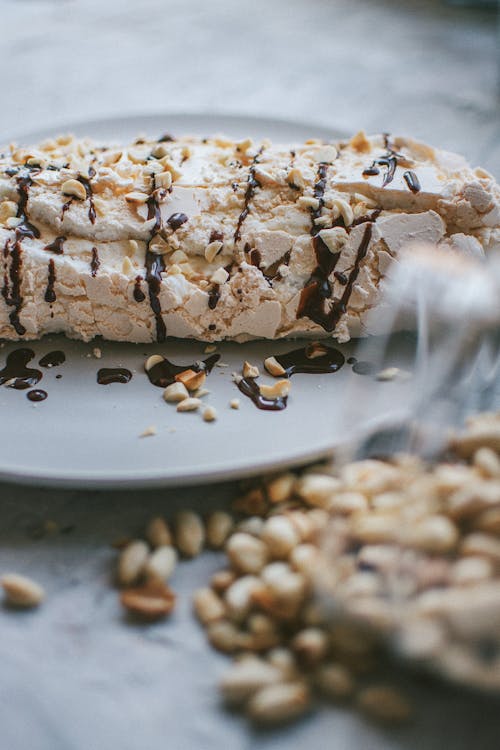 Free Sweet meringue roll with crust with chocolate topping served on plate near bag of pistachio nuts on table in kitchen Stock Photo