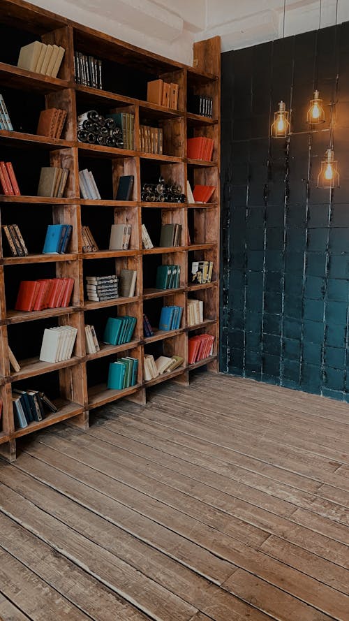 Wooden bookcase with collection of various books placed on wooden floor in light room with tiled wall and glowing light bulbs
