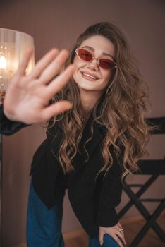 Smiling female with curly hair in trendy outfit and sunglasses looking at camera while standing in modern studio with chair