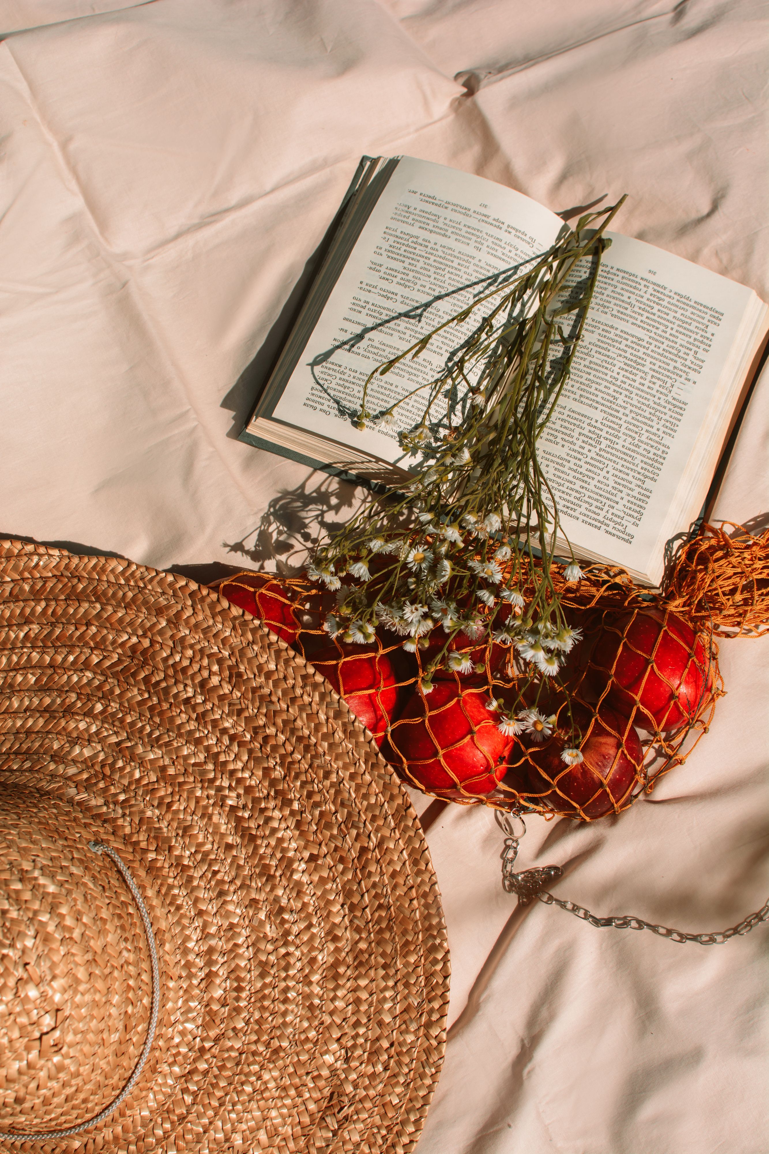 From above of straw hat placed near string bag with ripe apples near flower on opened book in sunny countryside
