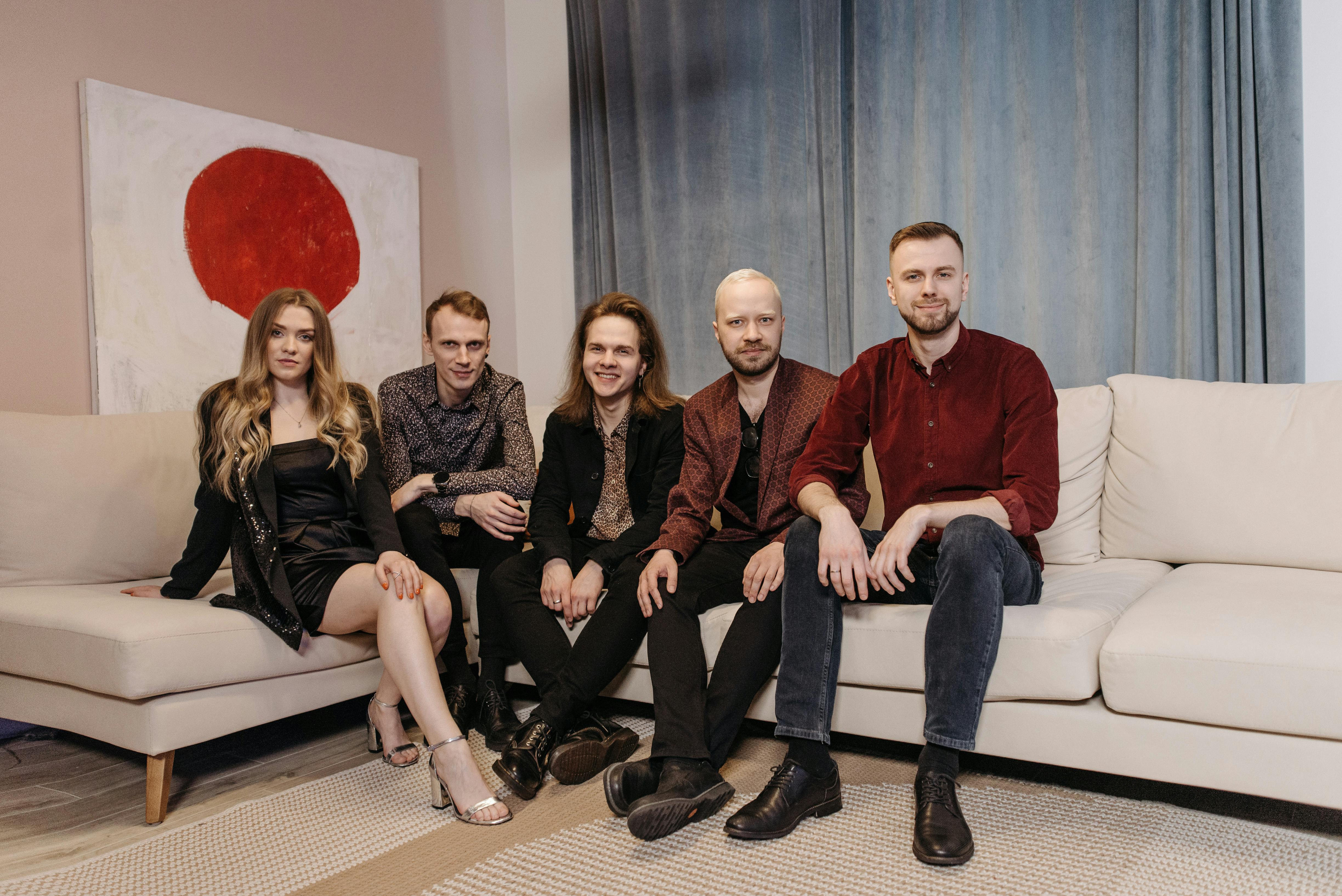 Five young adults sitting on a sofa indoors, smiling and relaxed.