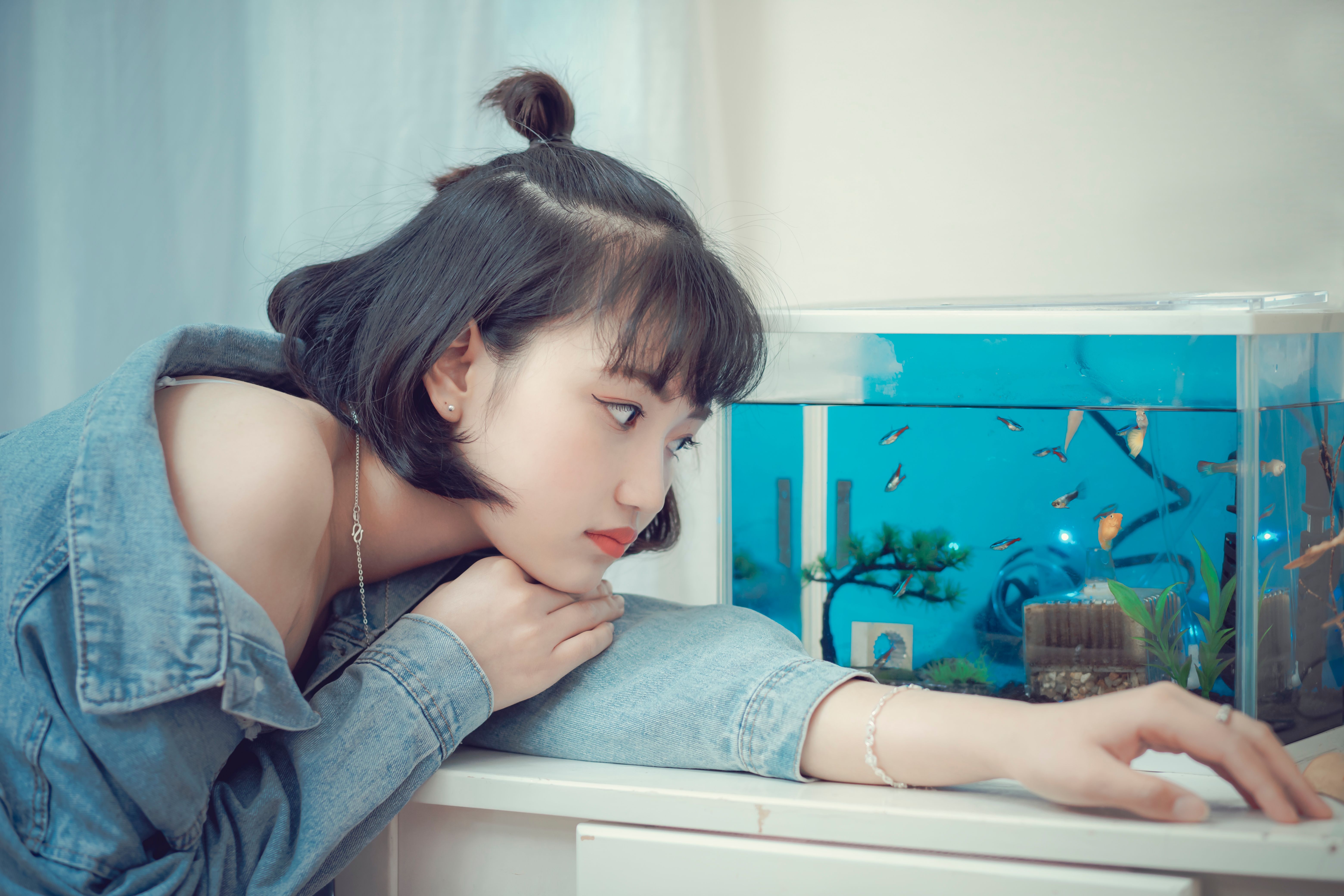 A young woman in a denim jacket observes fishes in a home aquarium.