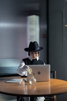 Elegant woman in a fedora hat using a laptop at a wooden table indoors. Modern style and focus.