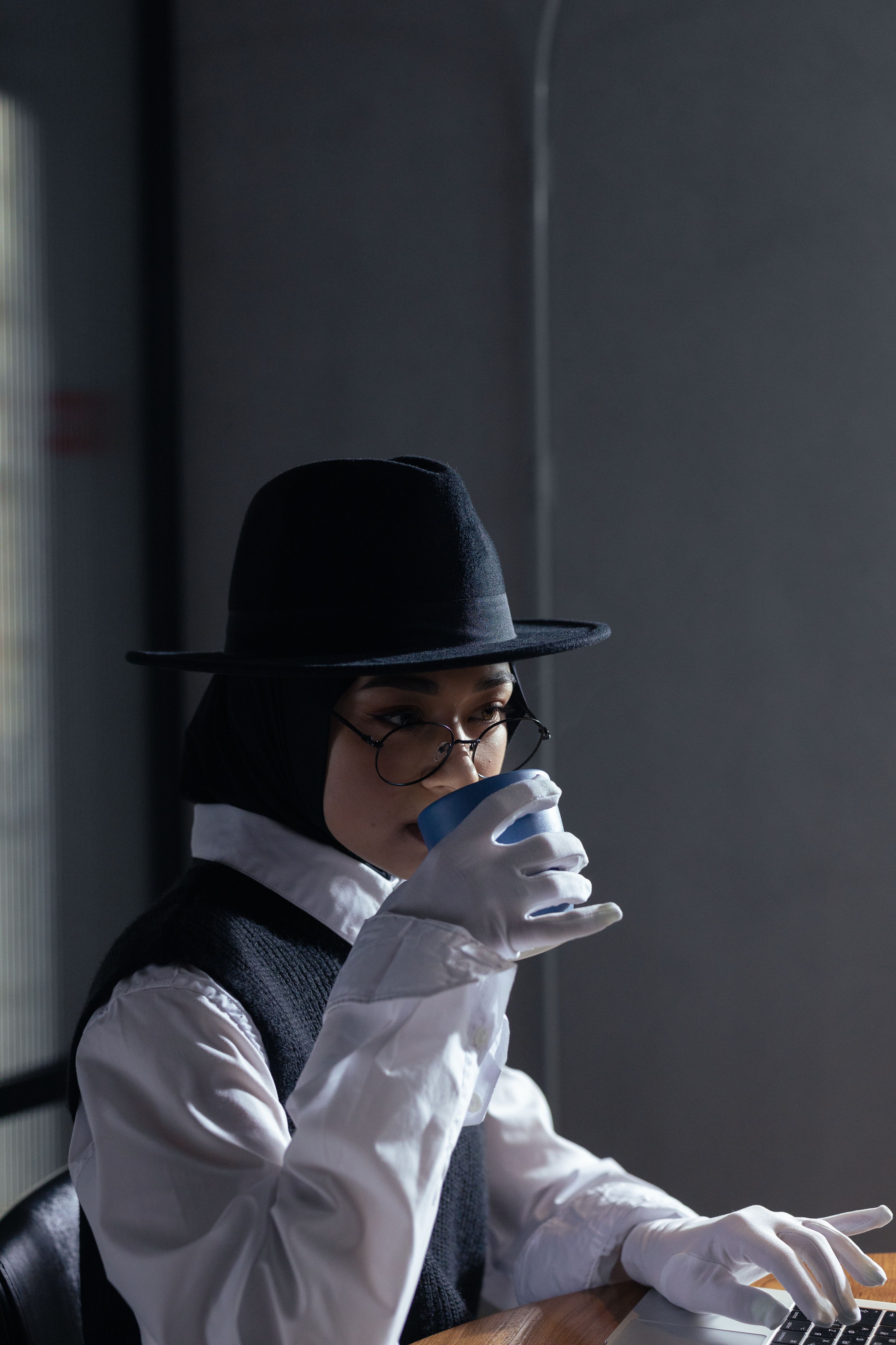 Mulher usando um chapéu fedora e óculos, bebendo café em um ambiente interno com estilo elegante.