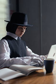 Woman in a fedora hat typing on a laptop, working indoors with a cup and open book nearby.