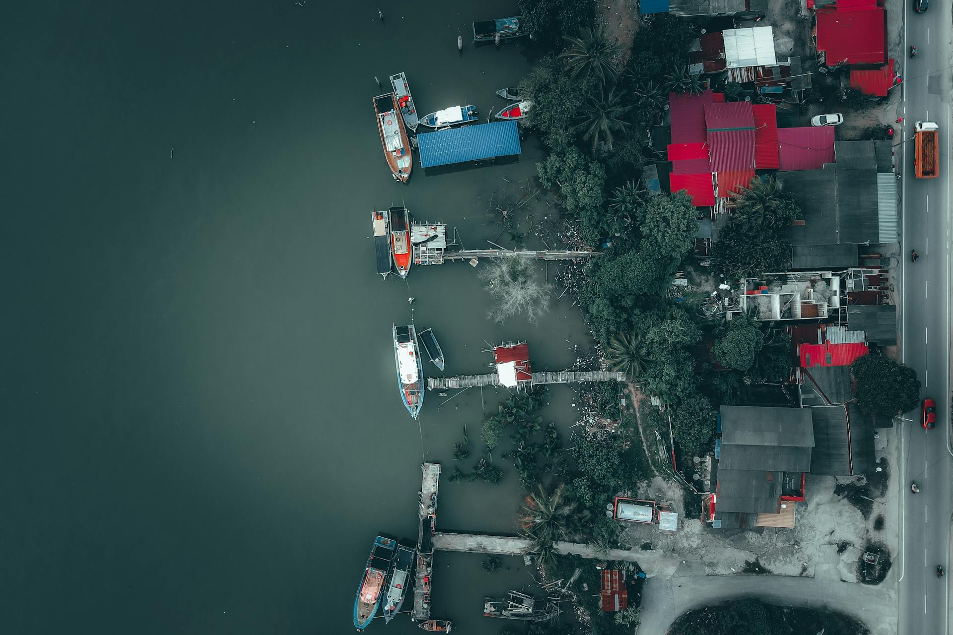 Aerial top view of boats moored in harbor near small houses surrounded by lush tropical trees near asphalt road