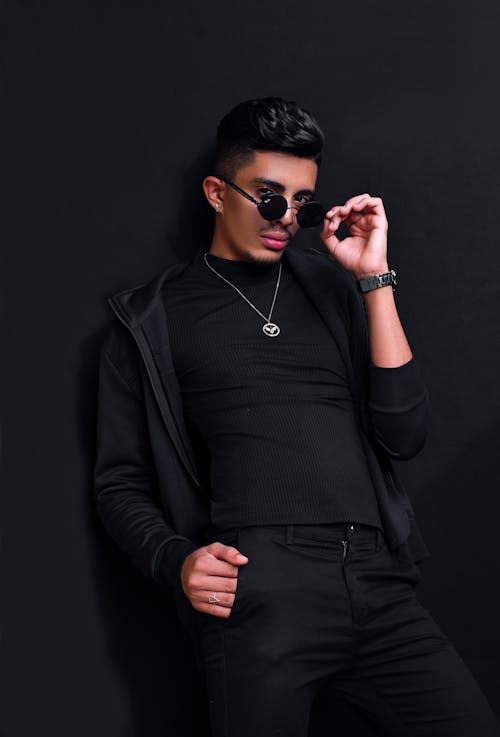Self confident young ethnic male model with dark hair in stylish outfit standing near black wall with hand in pocket and adjusting sunglasses in studio