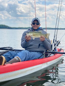 Man proudly holds fish while kayaking on a serene lake, wearing sunglasses and a gray jacket.
