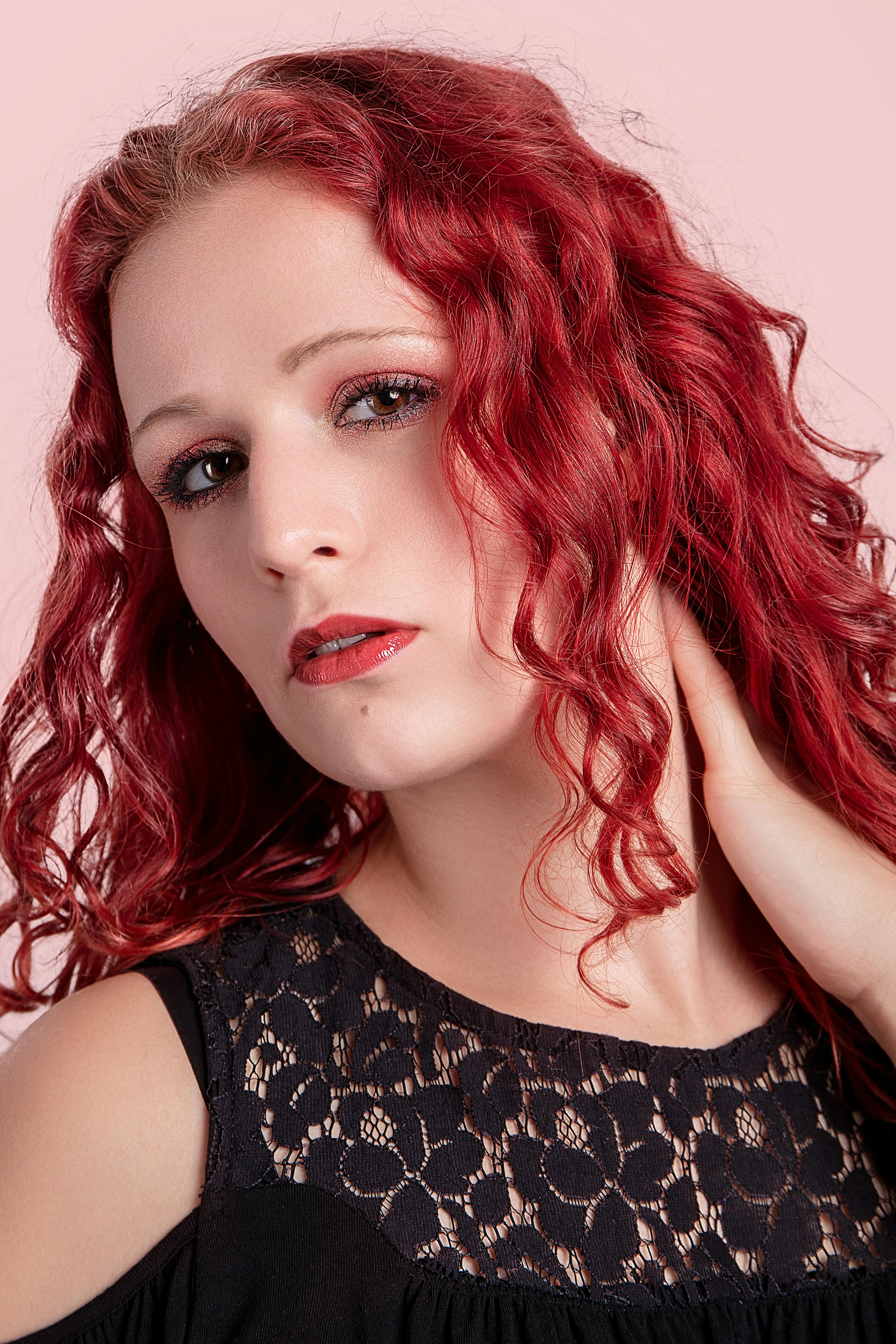 Close-up portrait of a woman with striking red curly hair against a pale pink background.