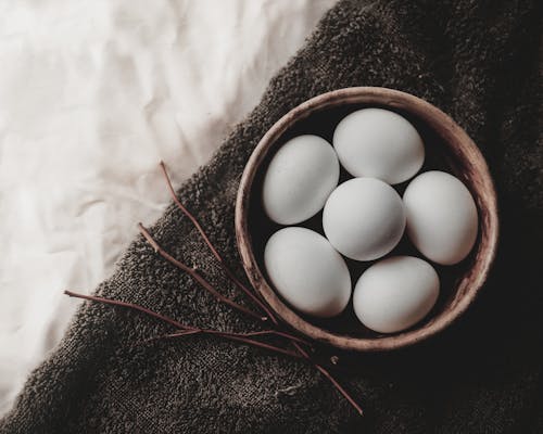 Top view of chicken eggs in wicker bowl placed near twigs on gray towel