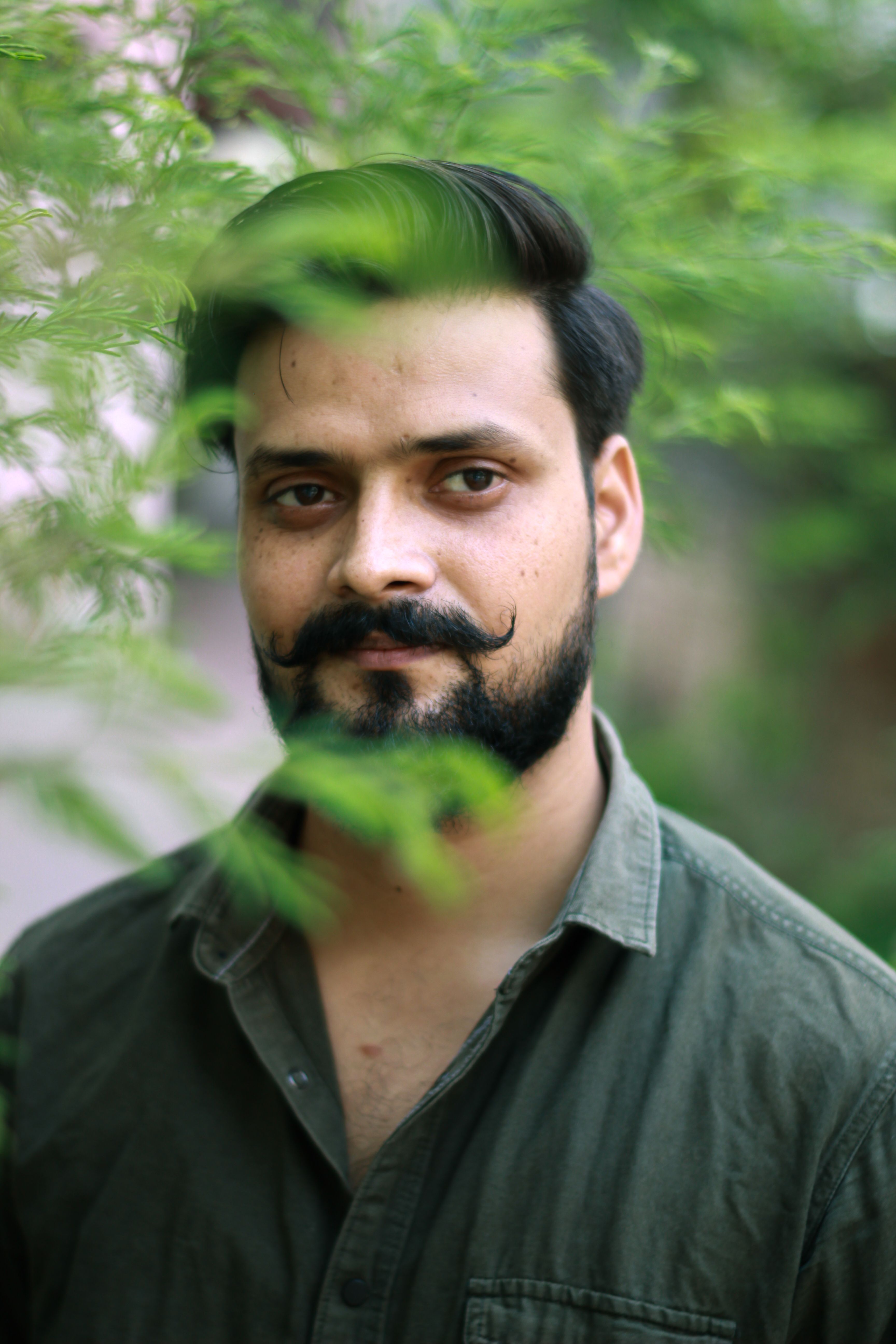 A man with a beard stands amidst green leaves, offering a serene and natural portrait setting.