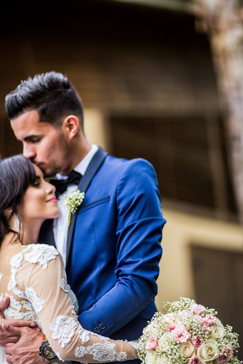 Young ethnic newlywed couple wearing elegant wedding clothes with bride bouquet hugging tenderly and kissing gently