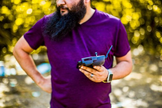 Crop unrecognizable male with beard in purple t shirt standing with smartphone with controller for drone in hand against blurred trees in sunny day
