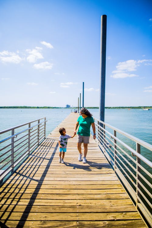 Female in casual clothes holding hand of child while walking on wooden pier above blue water of ocean in summer day