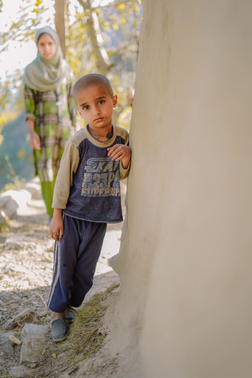 Free A young boy stands outdoors, leaning against a wall, with a distant woman in the background. Stock Photo