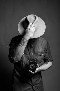 Black and white portrait of a man in a hat holding a vintage camera, exuding nostalgia.
