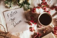 Top view of Follow Your Dreams inscription on paper placed on textile near mug of hot beverage and flower petals