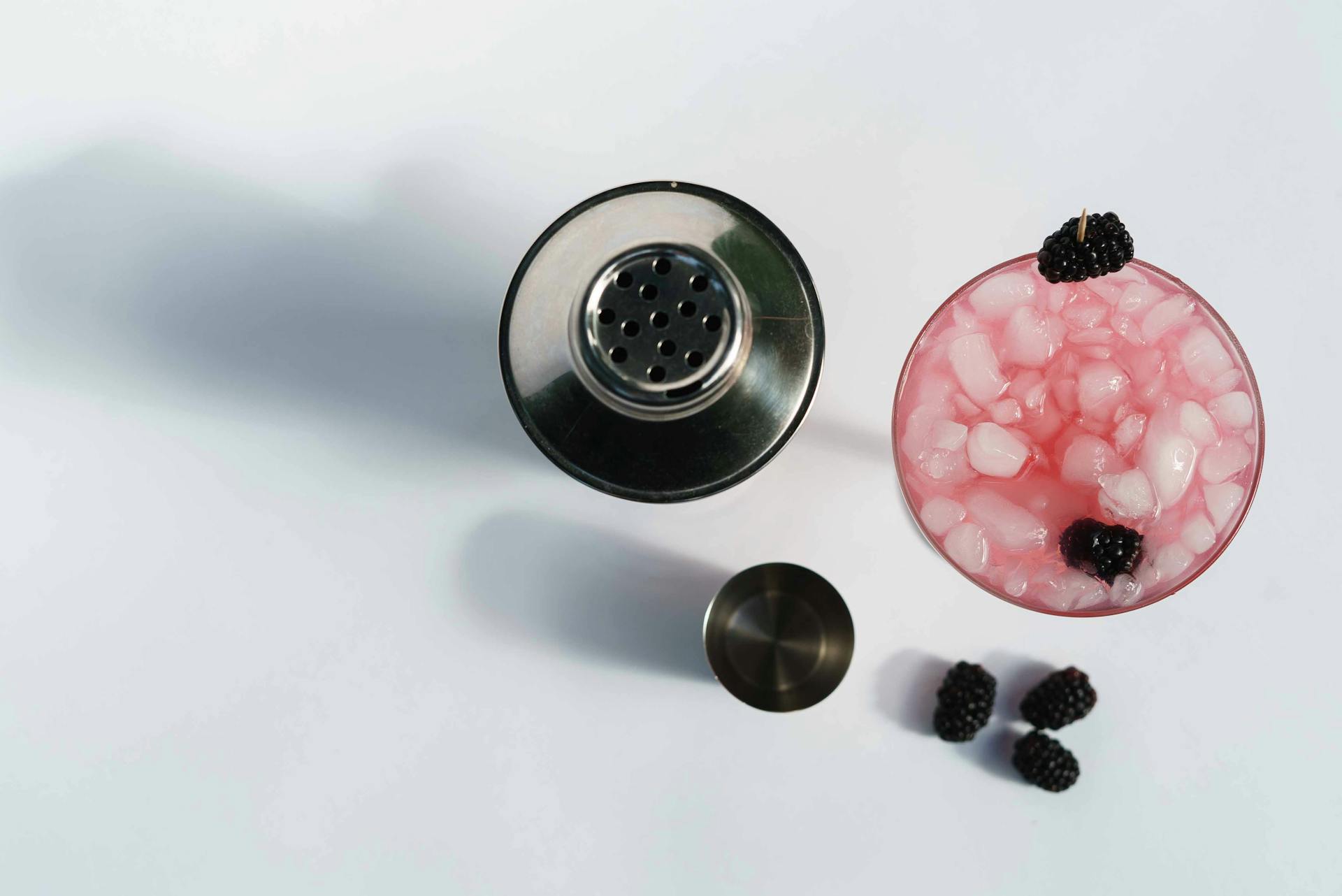 Top view of glass of cold blackberry margarita with berries and ice cubes near stainless shaker on white background