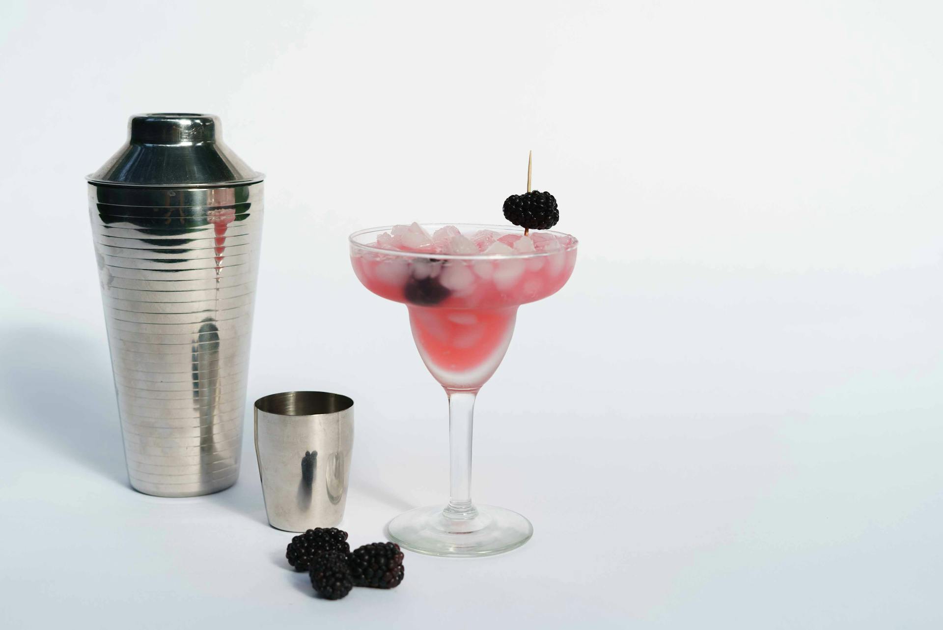 Cold refreshing margarita cocktail made of ice cubes and blackberries placed near shaker on white background