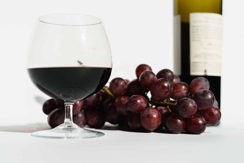Free Glass of red fragrant wine placed near bunch of sweet grapes and bottle in white studio Stock Photo