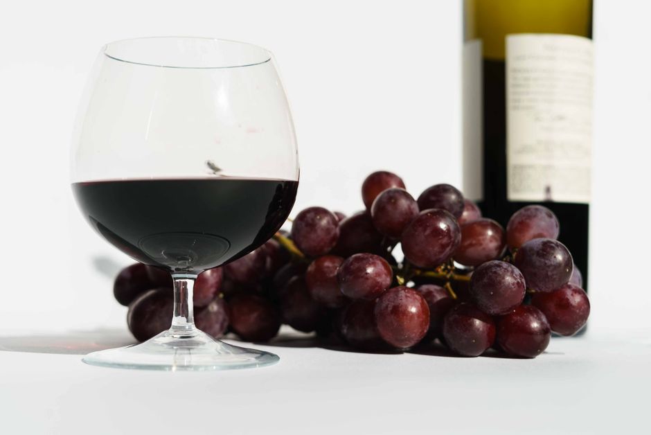 Glass of red fragrant wine placed near bunch of sweet grapes and bottle in white studio
