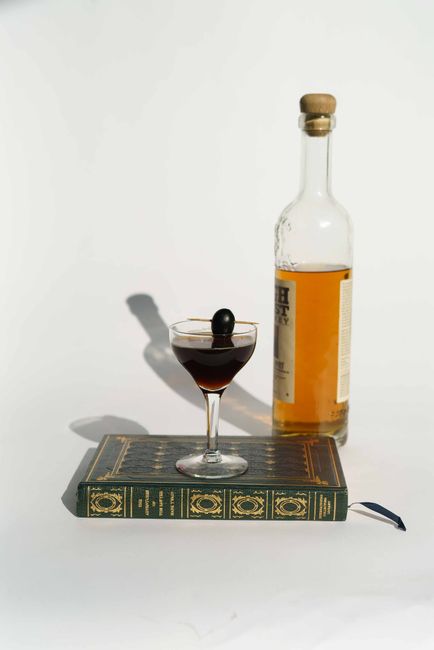 A Manhattan cocktail in a coupe glass, deep amber-red in color, garnished with a single brandied cherry on a pick.
