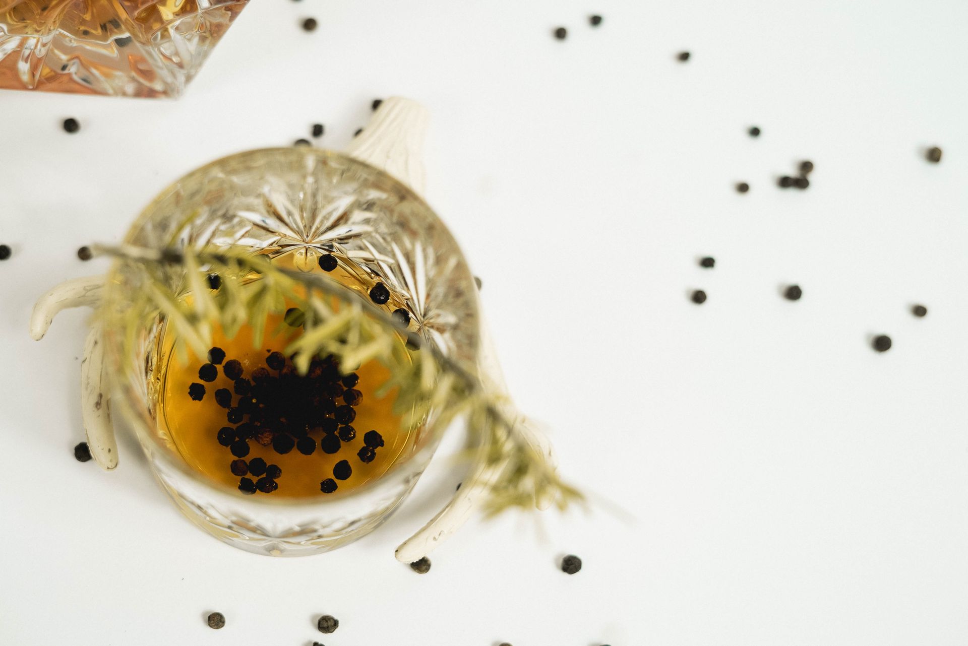 From above glass of whiskey with black peppercorns decorated with green rosemary twig placed on white background in light room