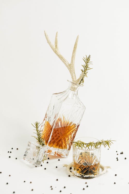Decanter with antlers and glass filled with whiskey decorated with rosemary twigs placed near scattered black peppercorns on white background