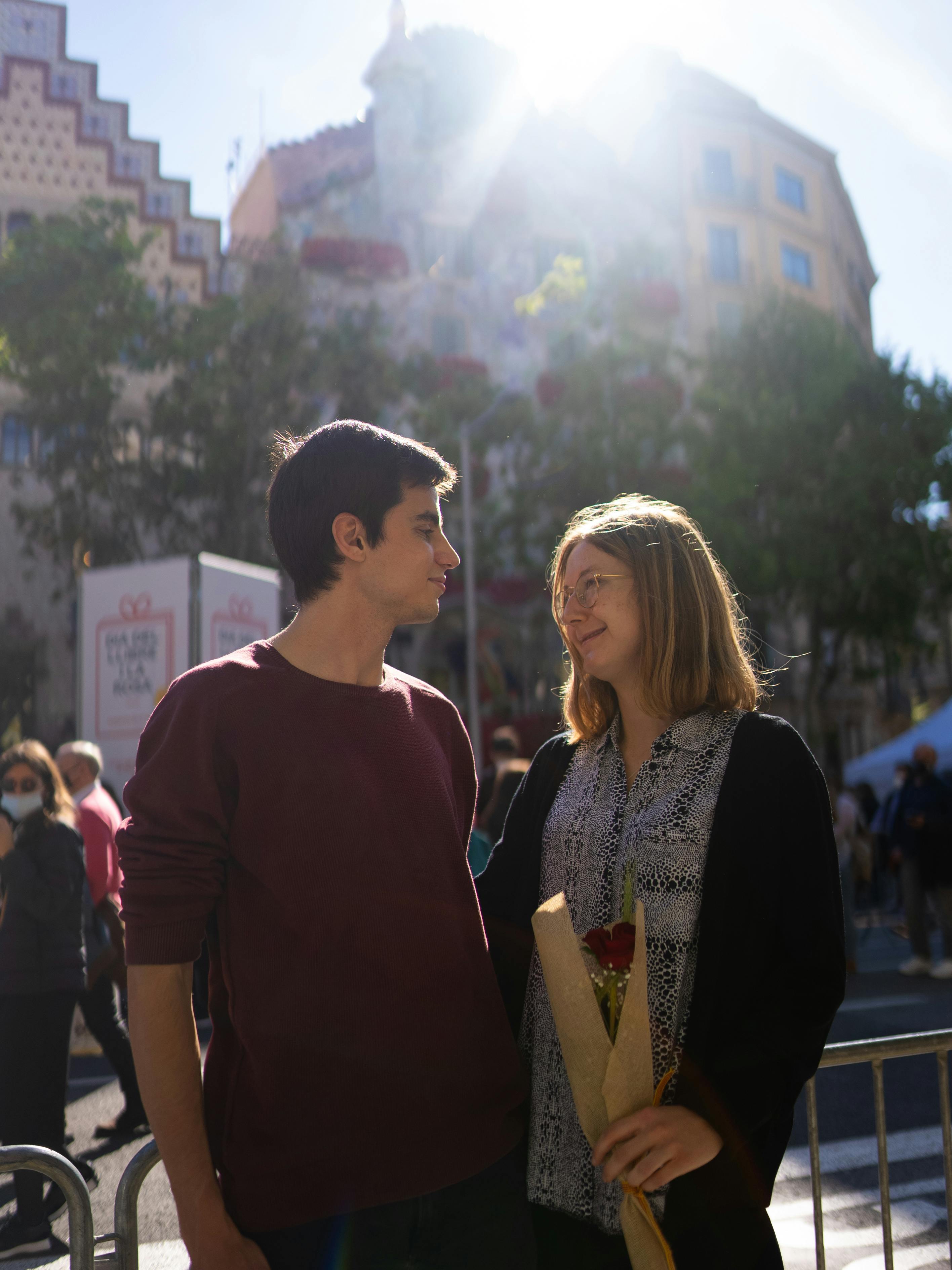 Smiling couple in Barcelona during a sunny festival holding a red rose bouquet, capturing love and happiness.